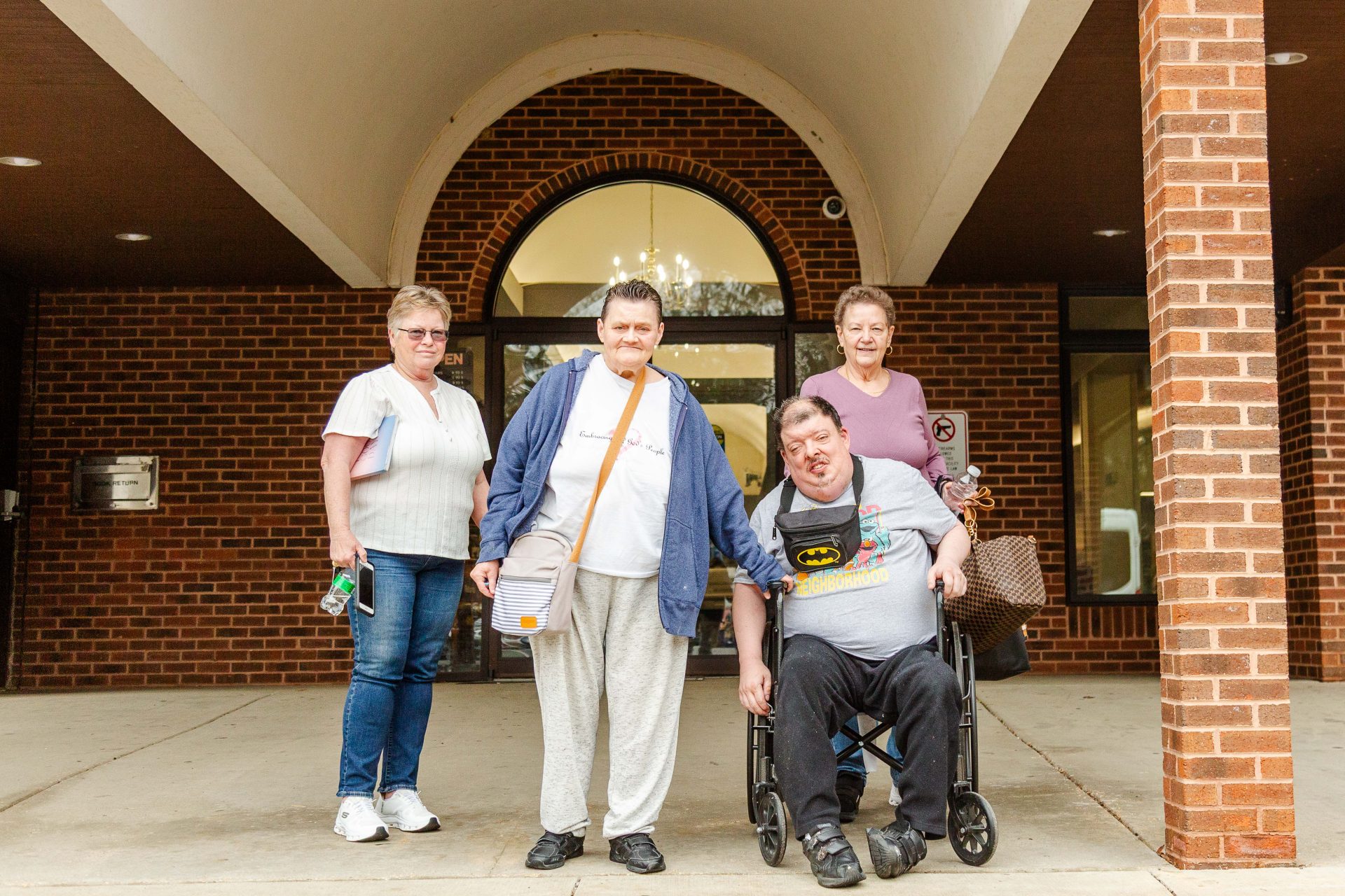 Four people, one in a wheelchair, in front of the entrance to a building. A brick column is visible in the foreground and windows in the background